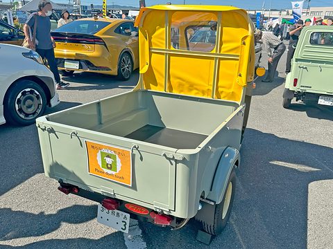 Rear of a pale green three-wheeled pickup with an empty bed, a yellow canvas cab and a ‘please don’t touch’ card. Opens a larger view
