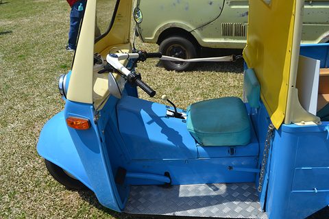 Open-sided three-wheeler cab with chrome handlebars, a turquoise seat cushion and a chequer-plate floor on grass. Opens a larger view