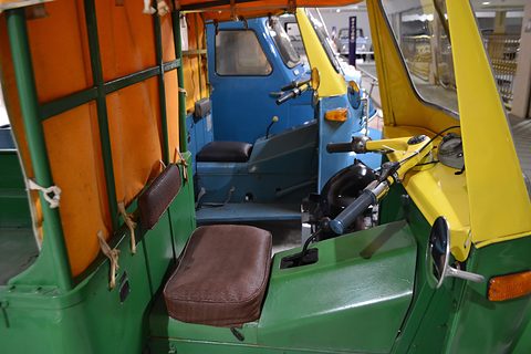 Inside a three-wheeler cab: a brown woven seat, yellow handlebars and orange canvas panels, a blue Midget behind. Opens a larger view