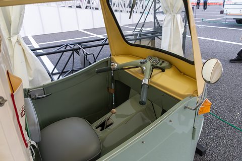 Open three-wheeler cab from the side: grey seat, handlebars with a small speedometer and a gear lever. Opens a larger view