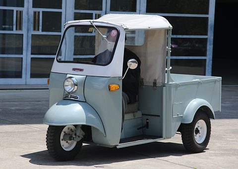 Pale grey-blue three-wheeled pickup with a white canvas top, one headlamp and a driver at the bars, on concrete. Opens a larger view