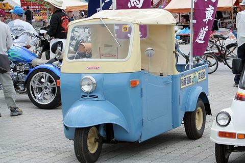 Light blue and cream three-wheeled pickup with lettering on the bed side, parked on paving among motorcycles and stalls. Opens a larger view