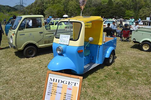 Blue and cream three-wheeled pickup on grass behind a framed MIDGET specification board, other old trucks around. Opens a larger view