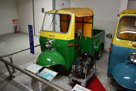 Green and yellow three-wheeled pickup with orange canvas cab sides, a bare engine on a trolley in front of it. Opens a larger view