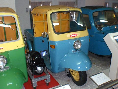 Blue three-wheeled pickup with a yellow cab between a green Midget at left and a blue truck at right, engine on a trolley. Opens a larger view