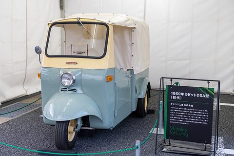 Pale green and cream three-wheeler with a canvas cab inside a white tent, beside a black information sign. Opens a larger view