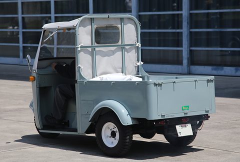 Rear three-quarter of a pale grey three-wheeled pickup with a white canvas cab and a drop-side bed on concrete. Opens a larger view