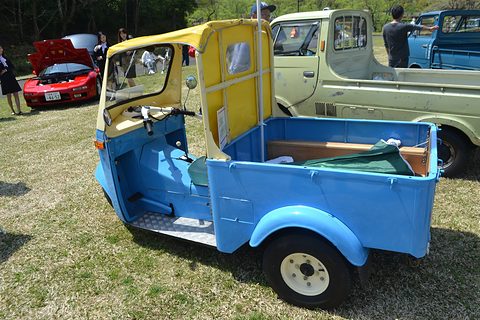 Blue three-wheeled pickup with a yellow cab and open load bed on grass, a red sports car behind. Opens a larger view