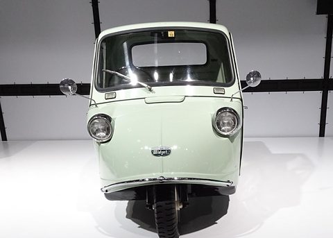 Head-on view of a pale green three-wheeled truck with two headlamps and one front wheel on a white stand. Opens a larger view