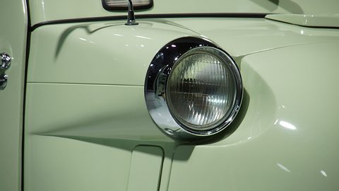 Close-up of a round headlamp with a chrome rim in a pale green curved wing, a side lamp above. Opens a larger view