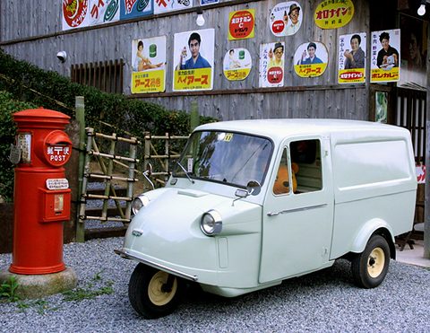 Pale green three-wheeled van with a closed box body beside a red cylindrical post box and old enamel signs. Opens a larger view