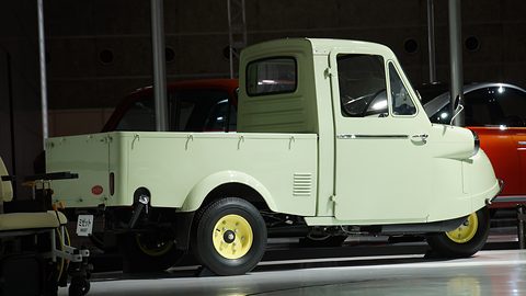 Side profile of a pale green three-wheeled pickup truck with a closed cab and drop-side bed on a white stand. Opens a larger view