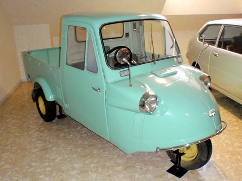 Turquoise three-wheeled pickup with a closed cab, chrome bumper bar and yellow wheels on a terrazzo floor. Opens a larger view