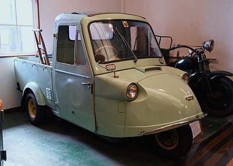 Weathered pale green three-wheeled pickup with a closed cab parked beside a black motorcycle by a window. Opens a larger view