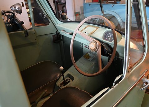 Pale green cab interior with a large brown two-spoke steering wheel, a floor lever and a dark vinyl bench. Opens a larger view
