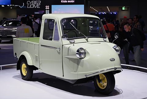 Pale green three-wheeled pickup with yellow wheels on a white turntable, show visitors behind.
