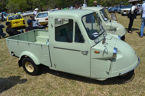 Pale green three-wheeled pickup with a closed cab on grass, a second Midget and visitors behind. Opens a larger view