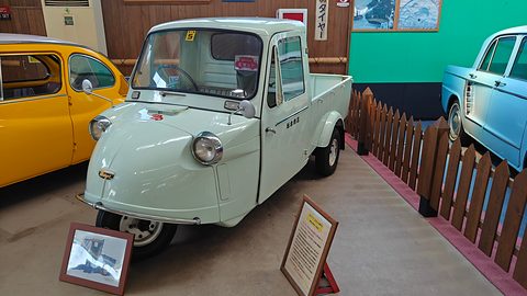 Pale green three-wheeled pickup beside a wooden fence with framed photographs and a sign in front. Opens a larger view