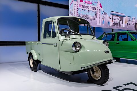 Pale green three-wheeled pickup with a chrome bumper bar on a white floor, a painted street scene and a green car behind. Opens a larger view