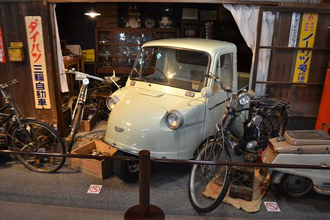 Pale green three-wheeled truck inside a recreated old shop with bicycles, a moped and a vertical Daihatsu sign. Opens a larger view
