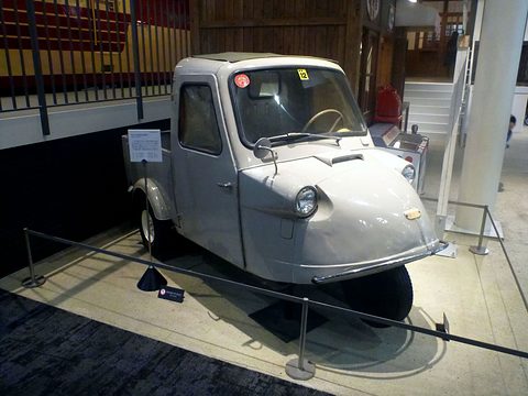 Grey three-wheeled pickup behind a low rope barrier in a museum hall with a railway carriage above. Opens a larger view