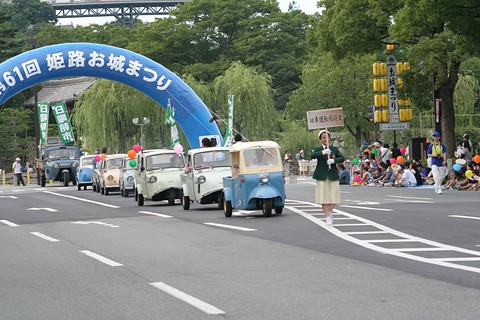 Line of small three-wheeled trucks driving along a wide road beneath a blue inflatable arch, spectators along the kerb.