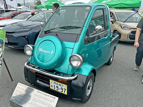Front of a turquoise tiny pickup with chrome bumper, round headlamps and a ‘Midget II’ spare-wheel cover. Opens a larger view