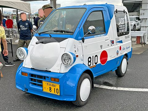 Blue and white tiny van with a red disc and number 810 on its door, visitors standing beside it. Opens a larger view