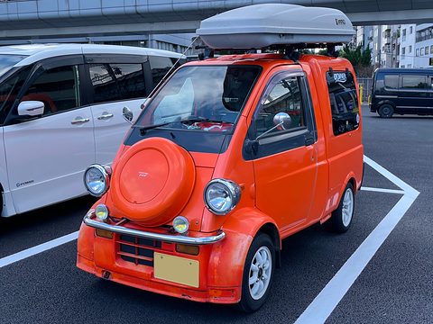 Orange tiny van with a round spare-wheel cover, chrome bumper and a grey roof box in a marked bay. Opens a larger view