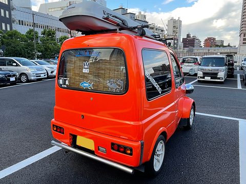 Rear of an orange tiny van with a bamboo blind in its rear window, a roof box and chrome bumper. Opens a larger view