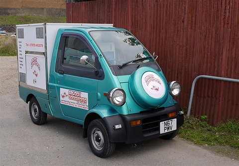 Teal tiny one-seat truck with a white sign-written box body and nose spare wheel beside a red wall. Opens a larger view