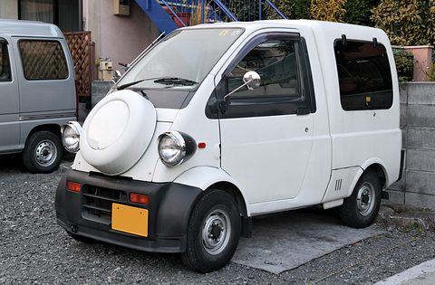 White tiny one-box van with a round spare-wheel cover on the nose and a yellow plate on gravel.