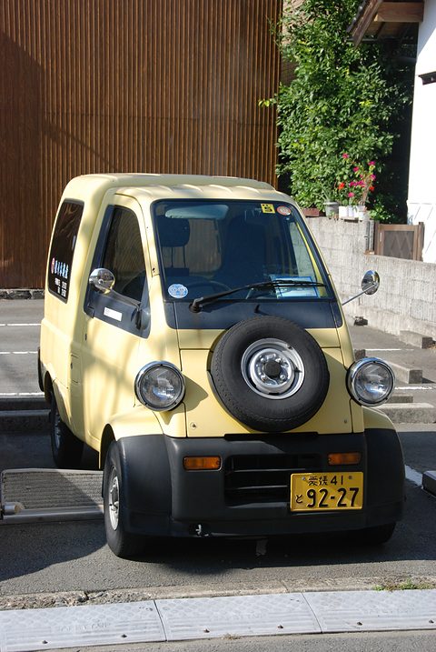Front of a cream tiny van with a black spare wheel on the nose and round headlamps by a wooden fence. Opens a larger view