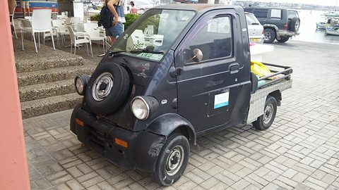 Dark blue tiny pickup with a nose-mounted spare wheel and round headlamps on paving by cafe steps. Opens a larger view