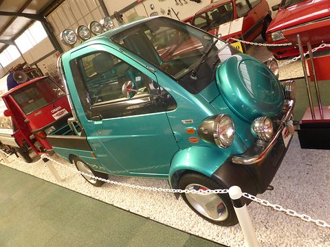 Teal tiny pickup with spotlights on its roof and a round nose cover, behind a chain on a museum floor. Opens a larger view
