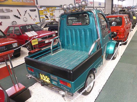 Rear of a teal tiny pickup with a row of chrome spotlights on its cab, red cars and wall displays around it. Opens a larger view
