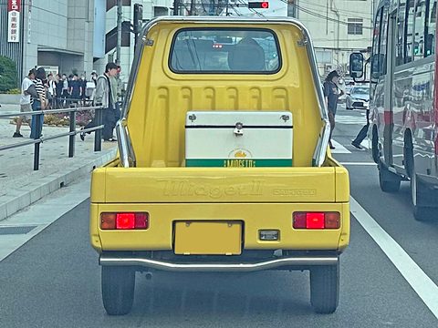 Rear of a yellow tiny pickup with a white cool box on its bed, driving in a city lane beside a bus. Opens a larger view