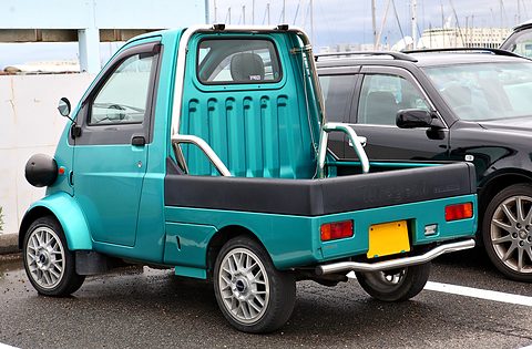 Rear of a turquoise tiny pickup with a chrome roll bar and alloy wheels beside a dark saloon. Opens a larger view