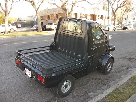 Rear of a black tiny pickup with a short ribbed cab back, a flat load bed and red tail lamps at a kerb. Opens a larger view