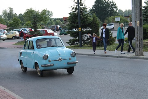 Pale blue small car driving on a wet road, a family walking along the pavement behind. Opens a larger view