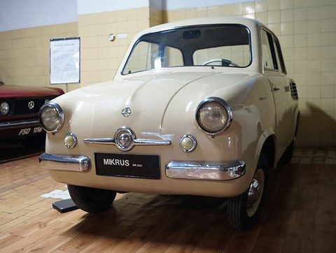 Cream small saloon with a MIKRUS name plate on a wooden floor before a tiled wall. Opens a larger view