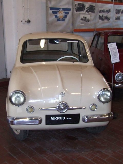 Cream small car facing the camera with a MIKRUS MR 300 plate, posters on the white wall behind. Opens a larger view