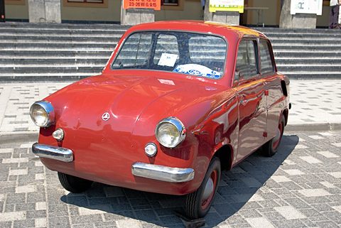 Red small saloon with chrome bumpers on paving before a flight of steps, a rally sticker in its windscreen. Opens a larger view