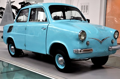 Sky-blue small two-door saloon with a chrome winged moustache on its nose, on a glass display floor. Opens a larger view