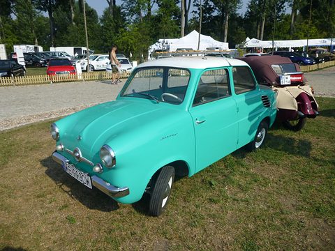 Turquoise small saloon with a white roof on grass, a cream and maroon microcar behind. Opens a larger view