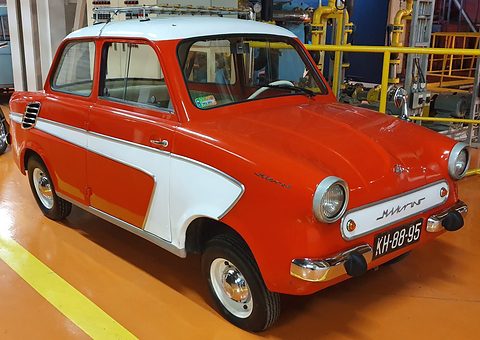 Red small saloon with white side flashes and a Mikrus nose plate in a factory hall with yellow railings. Opens a larger view
