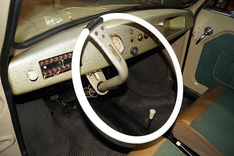 White two-spoke steering wheel before a speckled dashboard with a row of red-labelled switches, green seats. Opens a larger view