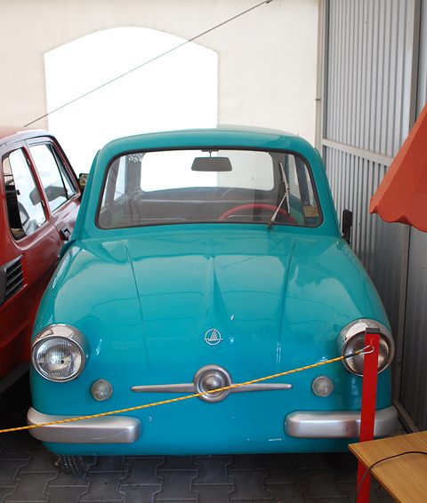 Turquoise small car head-on in a narrow shed behind a yellow rope, a red car beside it. Opens a larger view