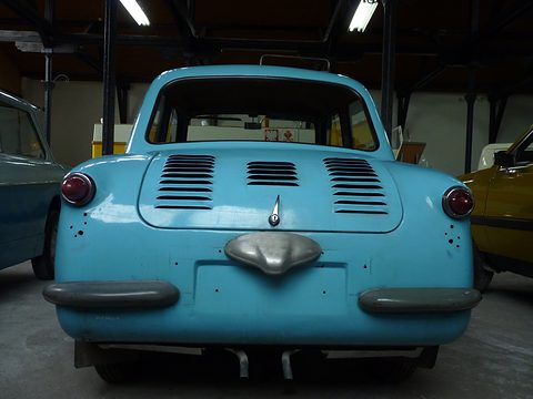 Rear of a sky-blue small car: louvred engine lid, chrome bumper guard and rubber overriders. Opens a larger view