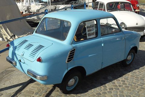 Pale blue small car from the rear on cobbles, boats and a white Syrena behind. Opens a larger view
